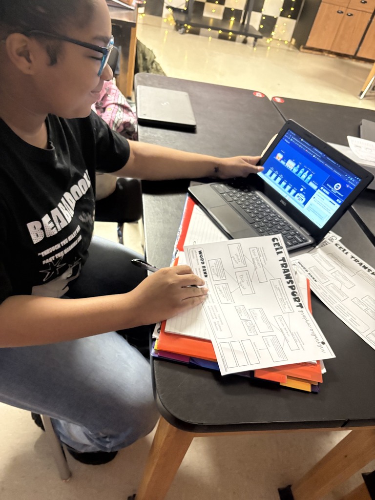 Student wearing glasses sitting at a table using a laptop and writing on a paper.