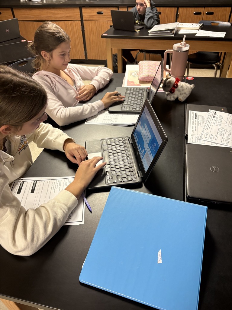 Two students sit at a table in a classroom, using laptops, with papers, pens, and folders on the table.