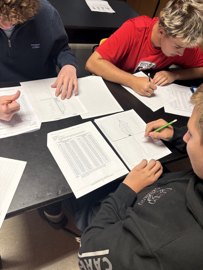 Three students seated at a table with papers and pens, writing on sheets of paper.