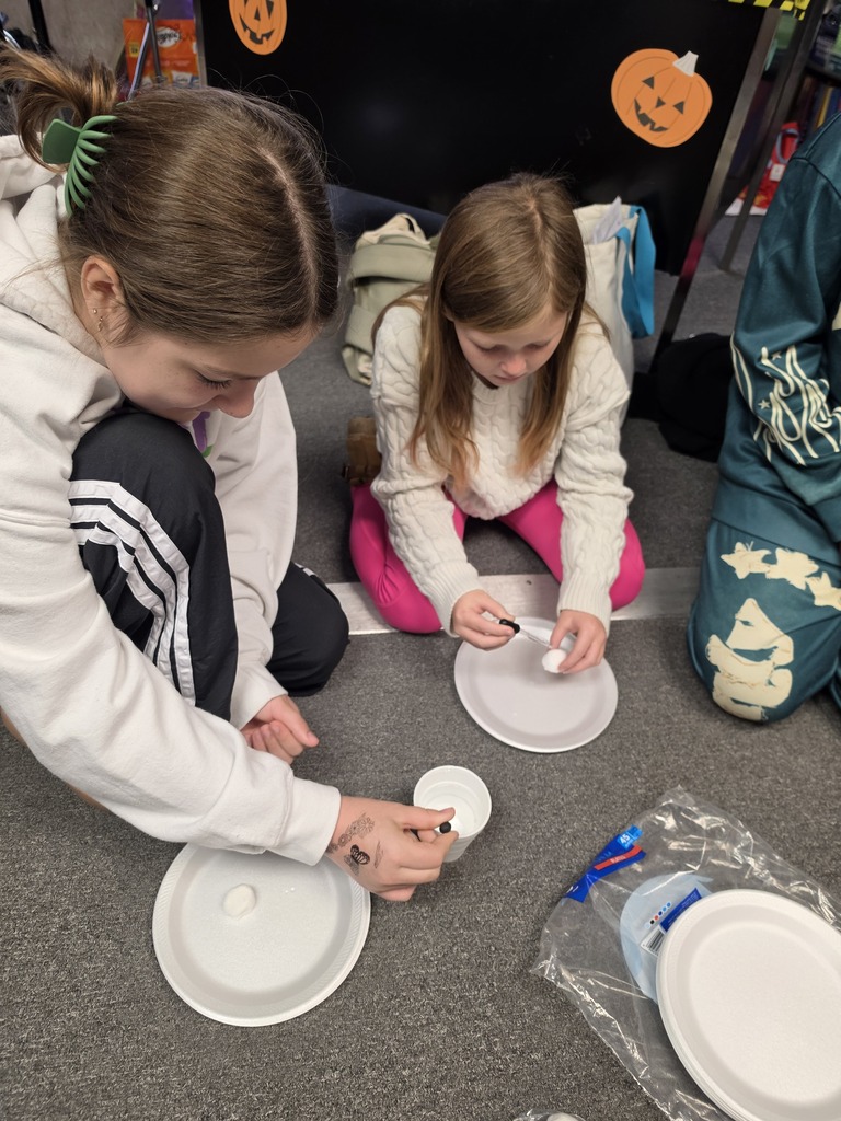 Two young girls are sitting on the floor. One is applying glue to a small object.