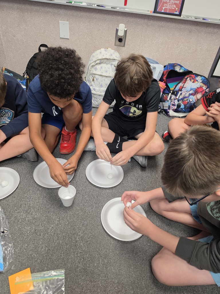 Four boys sit on the floor with plates and cups, preparing a science experiment.