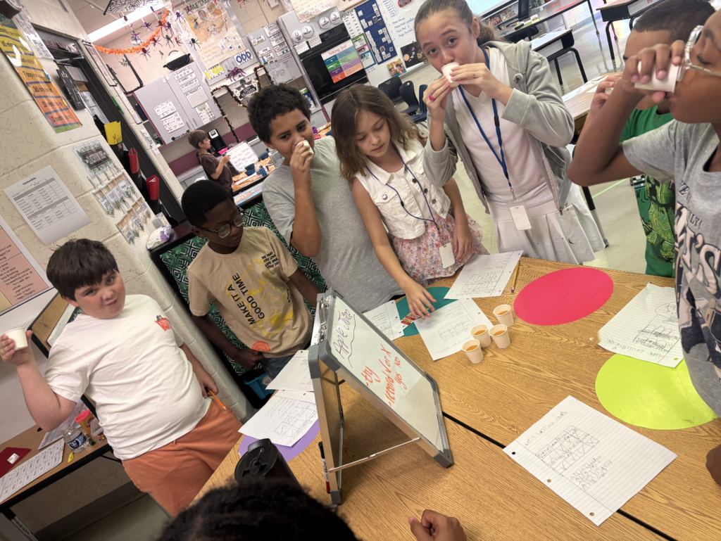 Children in a classroom taste food samples at a table, some holding cups. Whiteboard and papers are on the table.