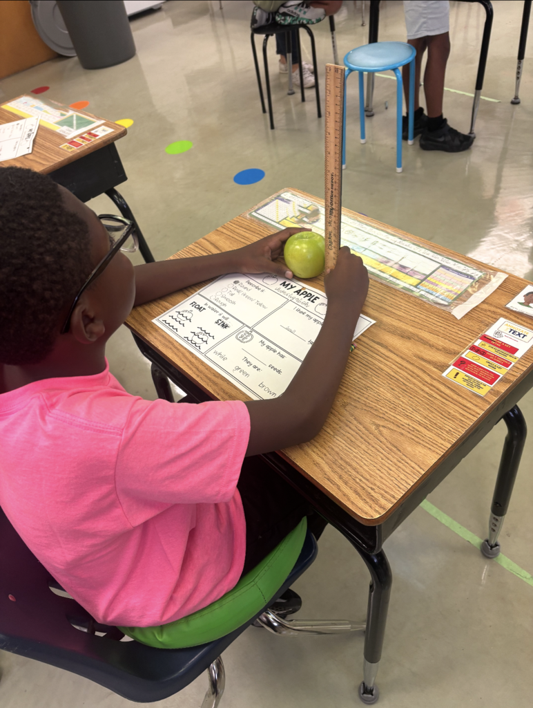 Child seated at desk, holding apple and ruler. Desk has papers, cards, and objects.
