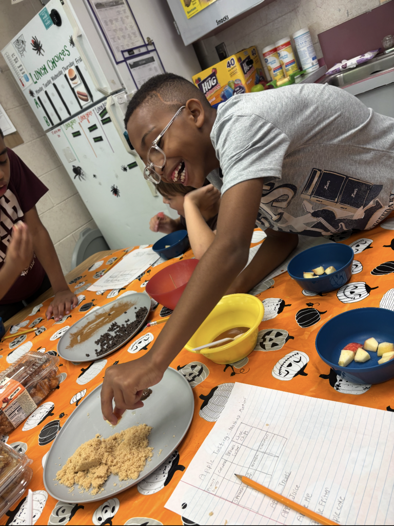 A boy mixes ingredients in a bowl at a table. Bowls, plates, and papers are on the table.