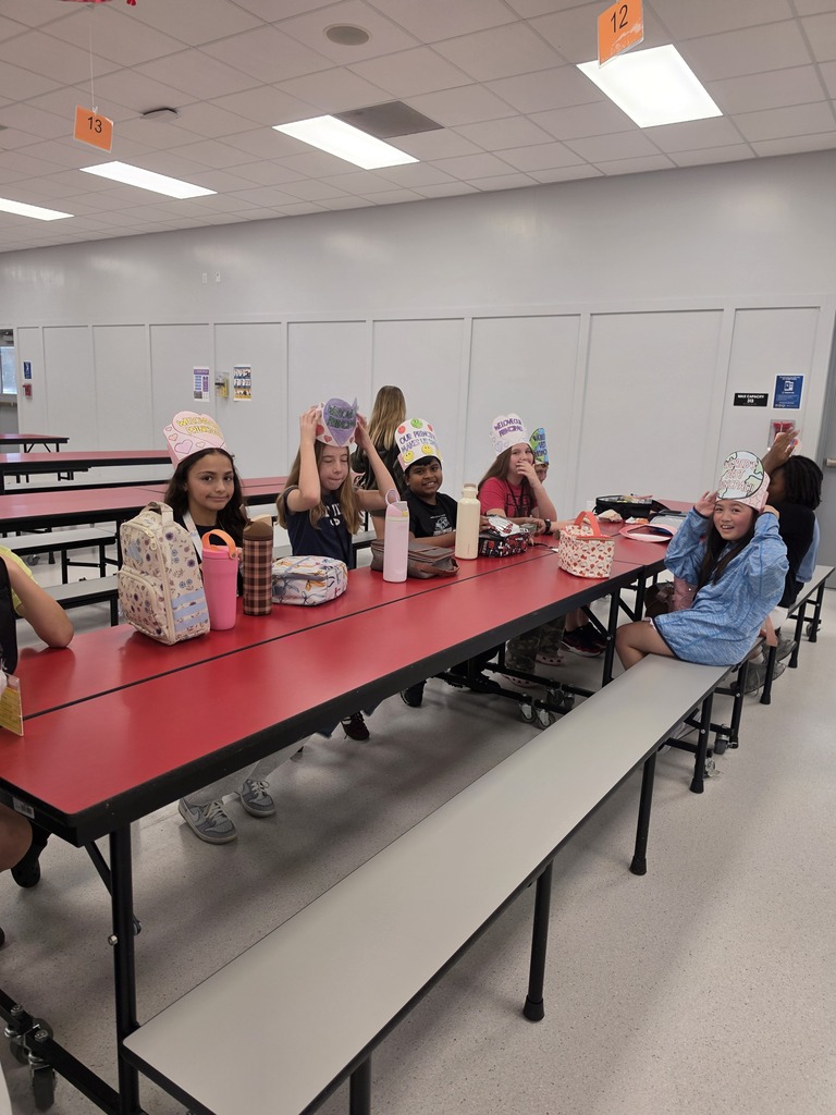 Students in a cafeteria wearing hats sitting at a long red table, with backpacks and cups.