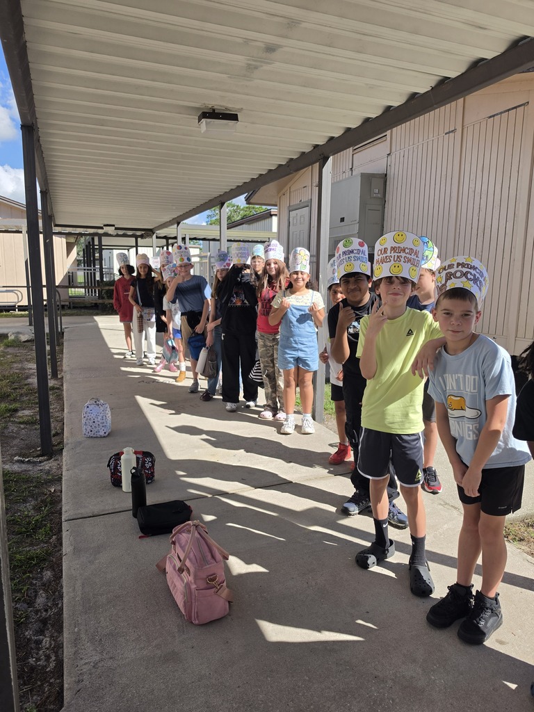 Several children wearing chef hats line up outside, with bags on the ground and a building in the background.