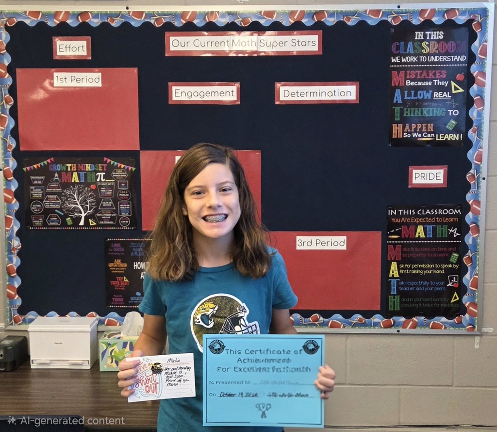A girl holds a certificate with blue and white text. She smiles and stands in front of a bulletin board.