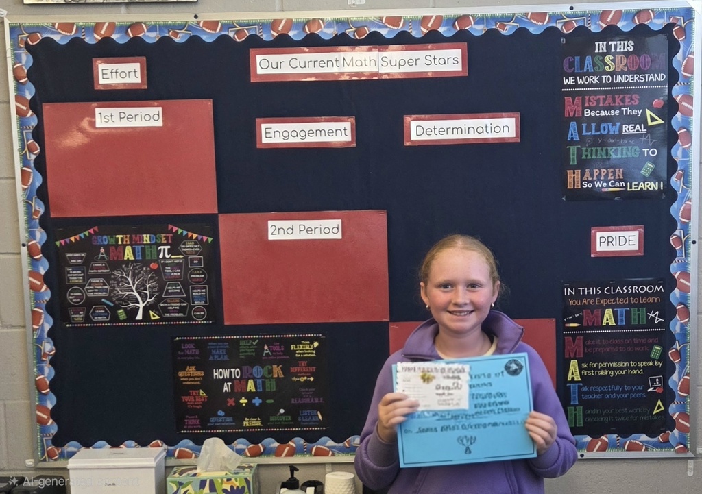 A young girl holding a blue certificate smiles in front of a colorful bulletin board with text.