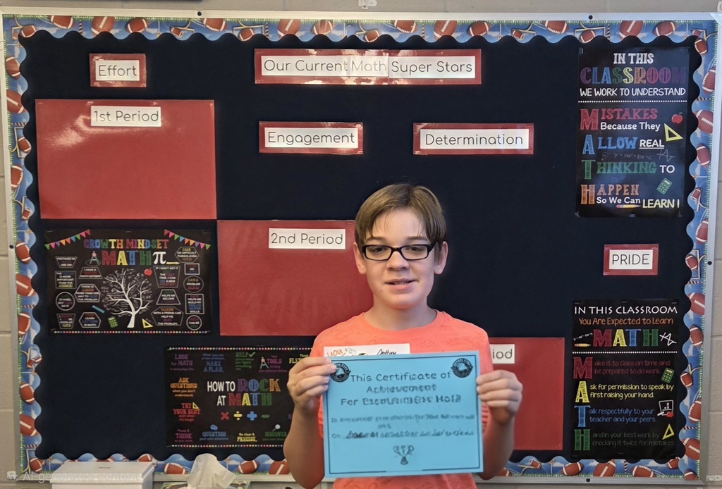 A student holds a certificate in front of a bulletin board with colorful squares and a tree design.