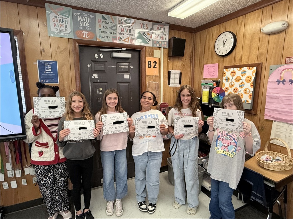 Six girls hold certificates in front of a classroom wall with a clock, bulletin board, and storage basket.