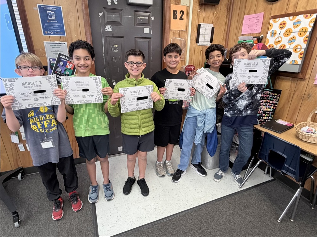 A group of students stand together, holding certificates, in a classroom with wooden walls and a table.