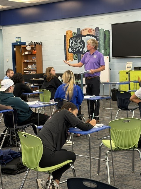 A classroom with students seated at desks, one seated person writing, and a man standing near a podium.