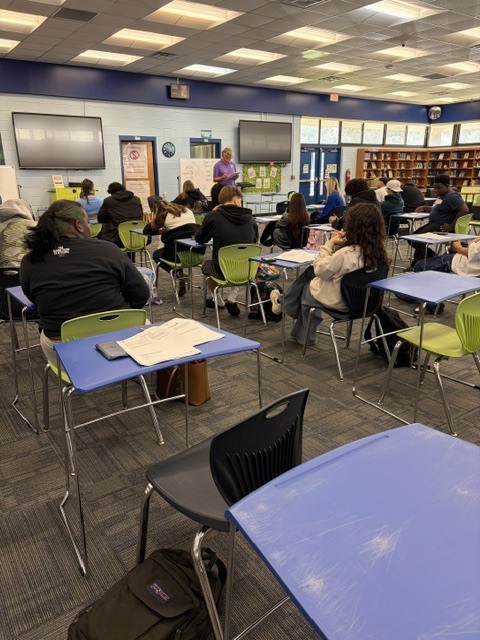 Classroom with desks and chairs. People sitting, some wearing jackets. A teacher at the front, with a whiteboard nearby.