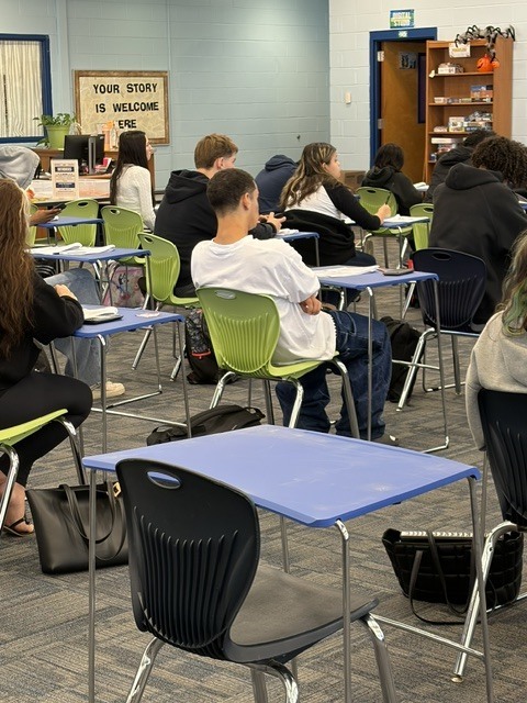 A classroom with students sitting at desks with backpacks; some sit with arms crossed.