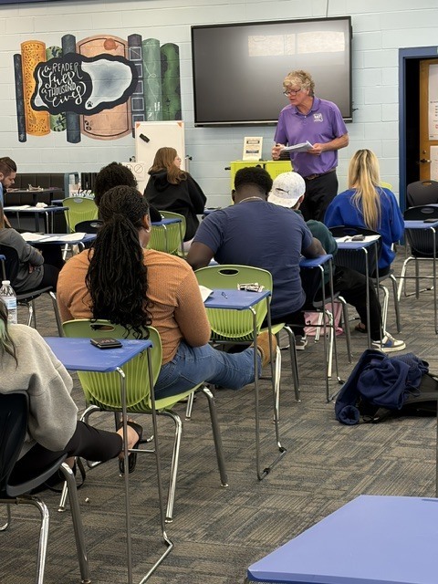 Classroom with chairs and tables, people seated, one person standing at a podium, a monitor on the wall.