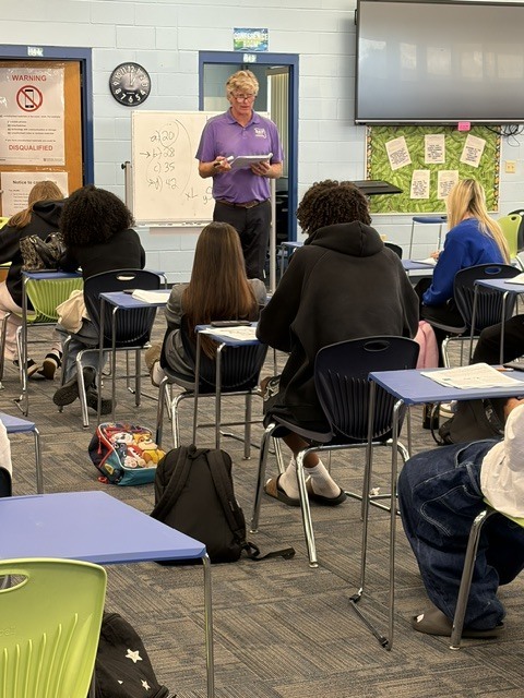 A classroom with desks and chairs, a man in purple stands at a podium with a whiteboard.