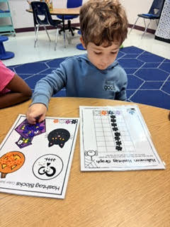 Boy sits at table, studying worksheet with pumpkins, cats, and bats, another worksheet with number grid.