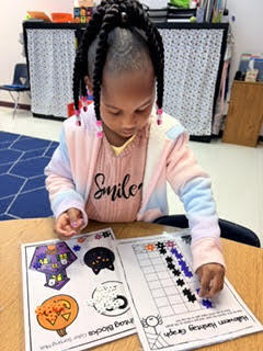Girl wearing pink jacket seated at a table working on a worksheet with fall theme.