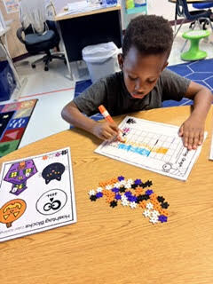Student sits at desk, drawing and coloring a worksheet with a marker. Multi-colored beads on desk.