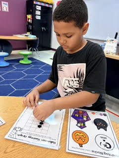Child sits at desk, working on a paper with fall images. The boy is wearing a black t-shirt.