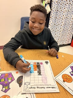 Child with dark hair smiles while coloring a worksheet. Table holds a worksheet with fall-themed drawings and colored markers.