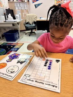 Child seated at a desk, working on a worksheet with a grid and various fall-themed images.