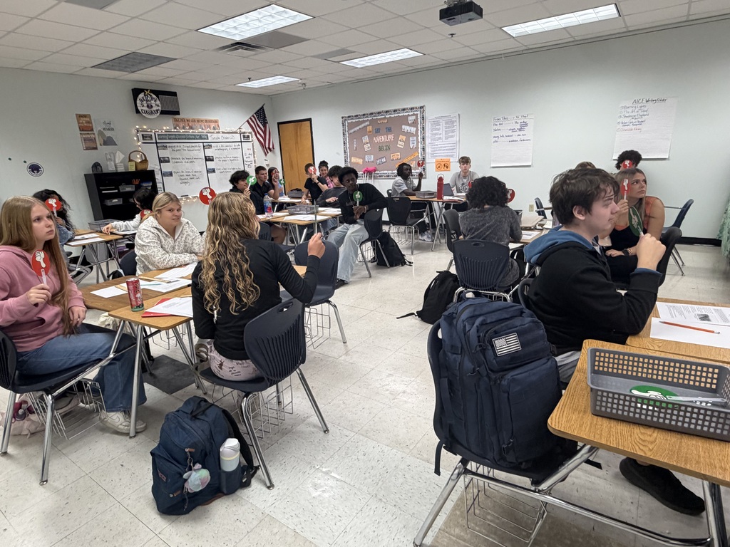Students in a classroom sit at tables, engaged in discussions, with backpacks and papers visible.