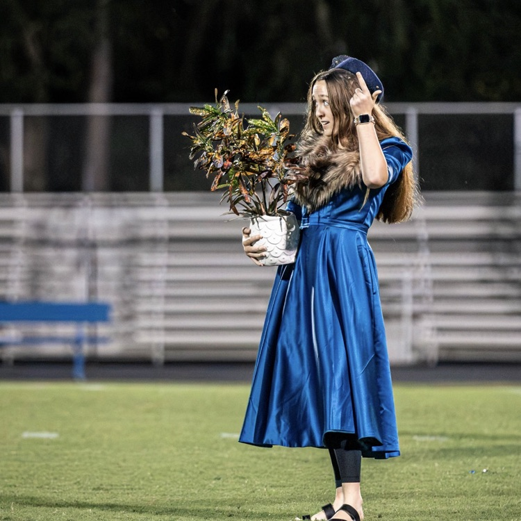 Woman in blue dress holding plant in pot stands on field at night; bleachers behind.