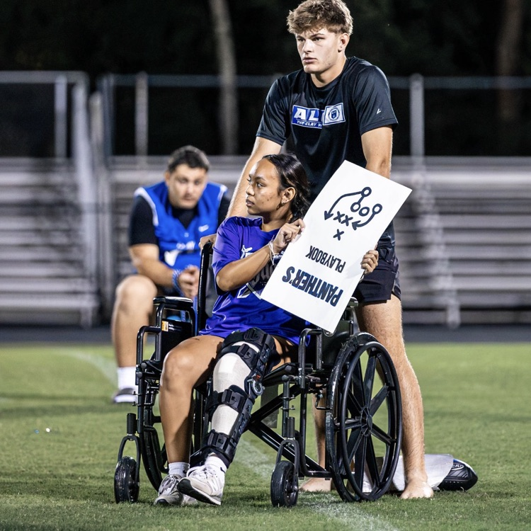 Two young adults with one in a wheelchair holding a sign on a field with bleachers in the background.