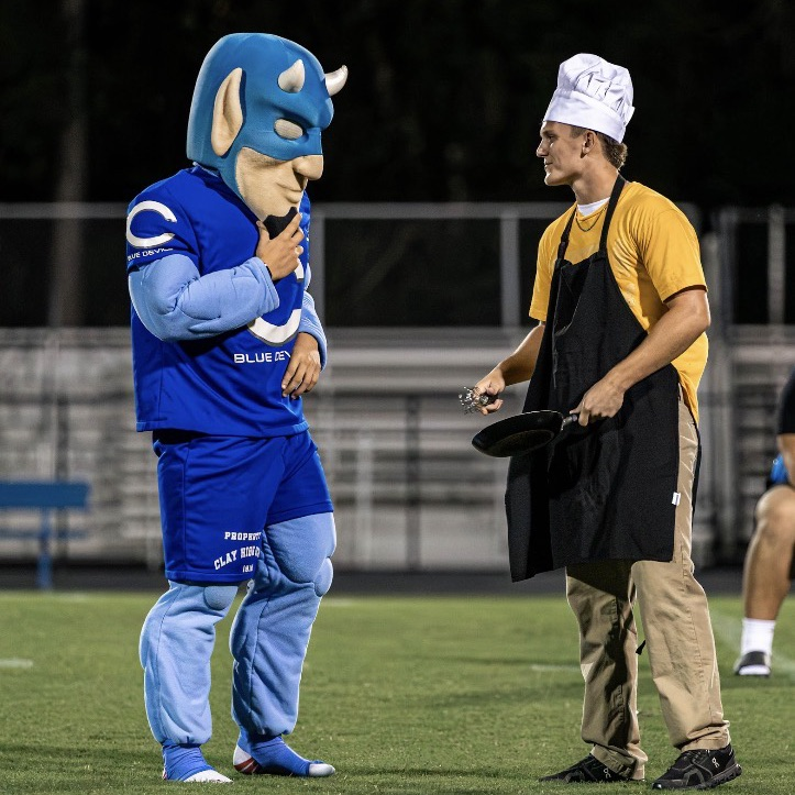 A mascot wearing blue and a man in a yellow shirt and black apron stand on a field.