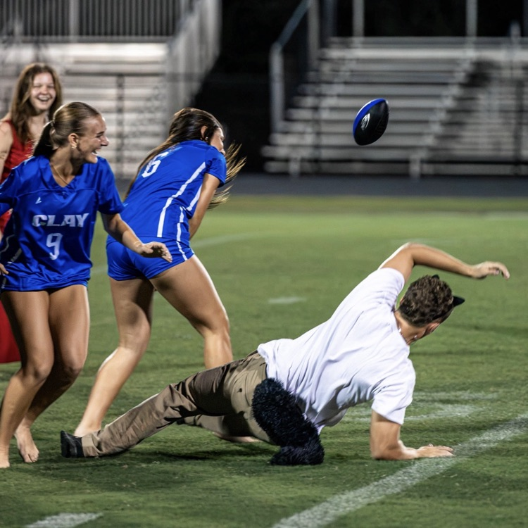 A football player falls to the ground on a field as three women look on, with bleachers in the background.
