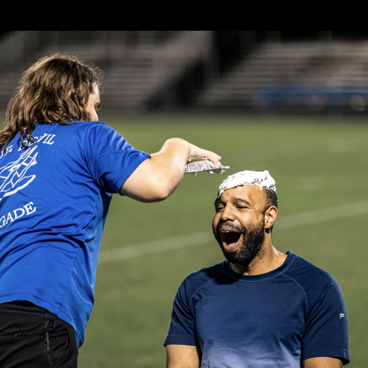 Woman in blue shirt holds white object, placing it on man's head; both look at camera. Background includes stadium bleachers.