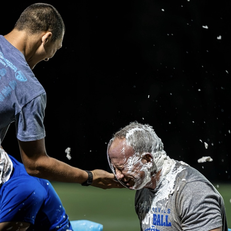 Two men sitting in blue chairs with a third man applying white substance to the face of one.