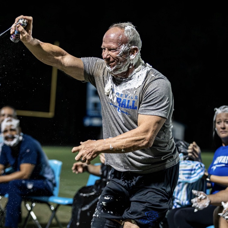 Person in gray shirt holding whipped cream container at outdoor sports event, pointing while people sit in chairs.