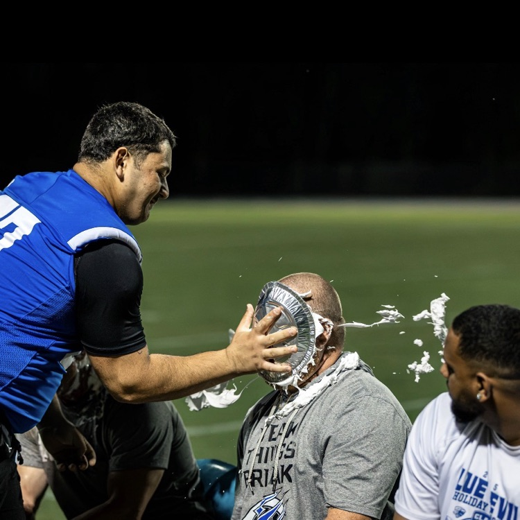 Two men in jerseys pour liquid over the head of a seated man. Other people in jerseys stand nearby.