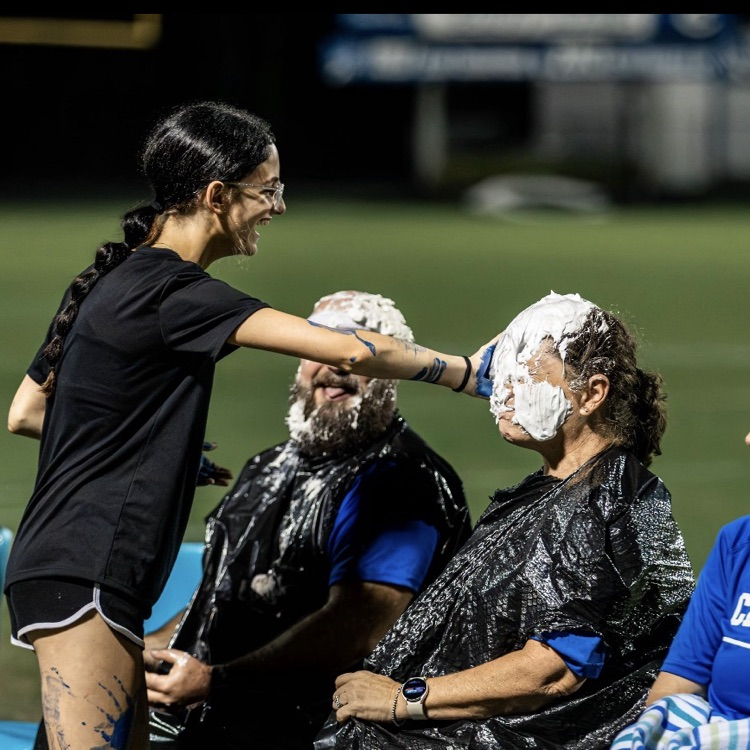 Woman with braided hair smears white substance on another woman's face on a sports field.