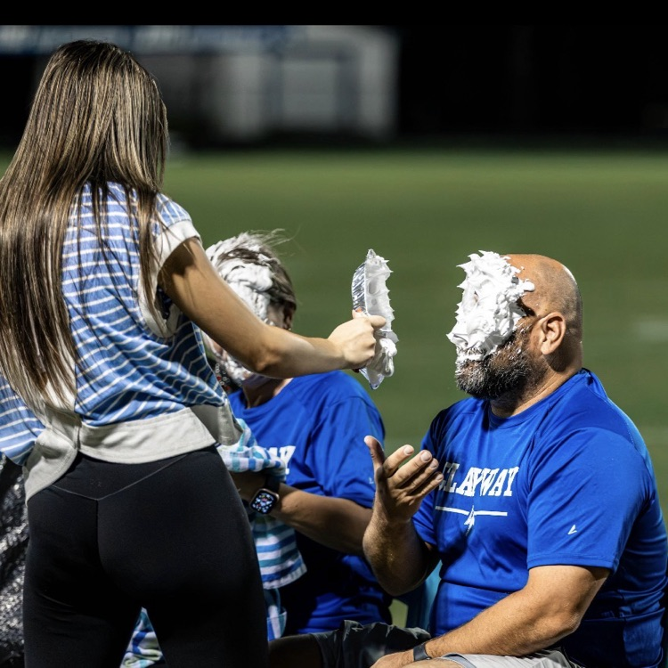 A person with a face covered in white cream sits in a chair at a football game.