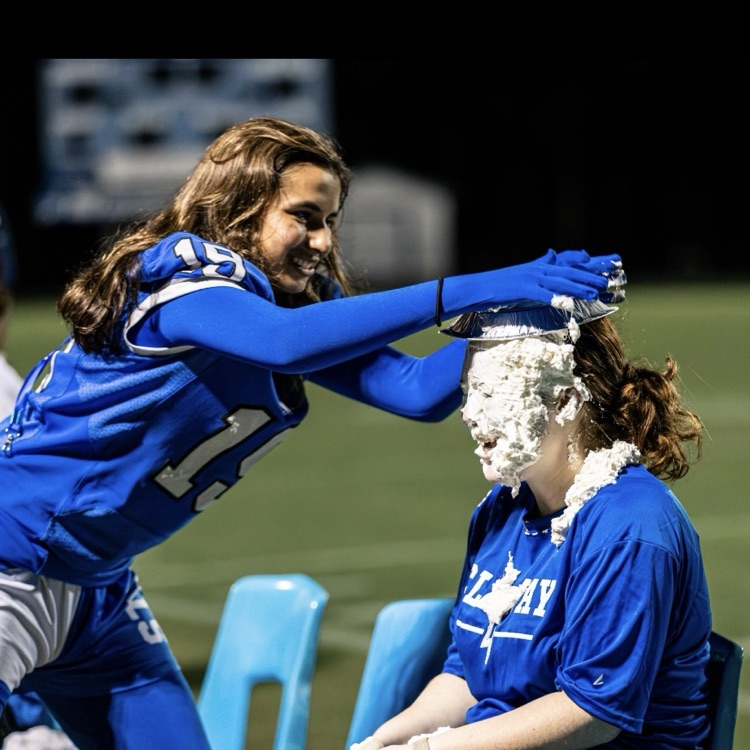 Two people, one in blue, apply substance to a woman's head. She sits in a chair on a football field.