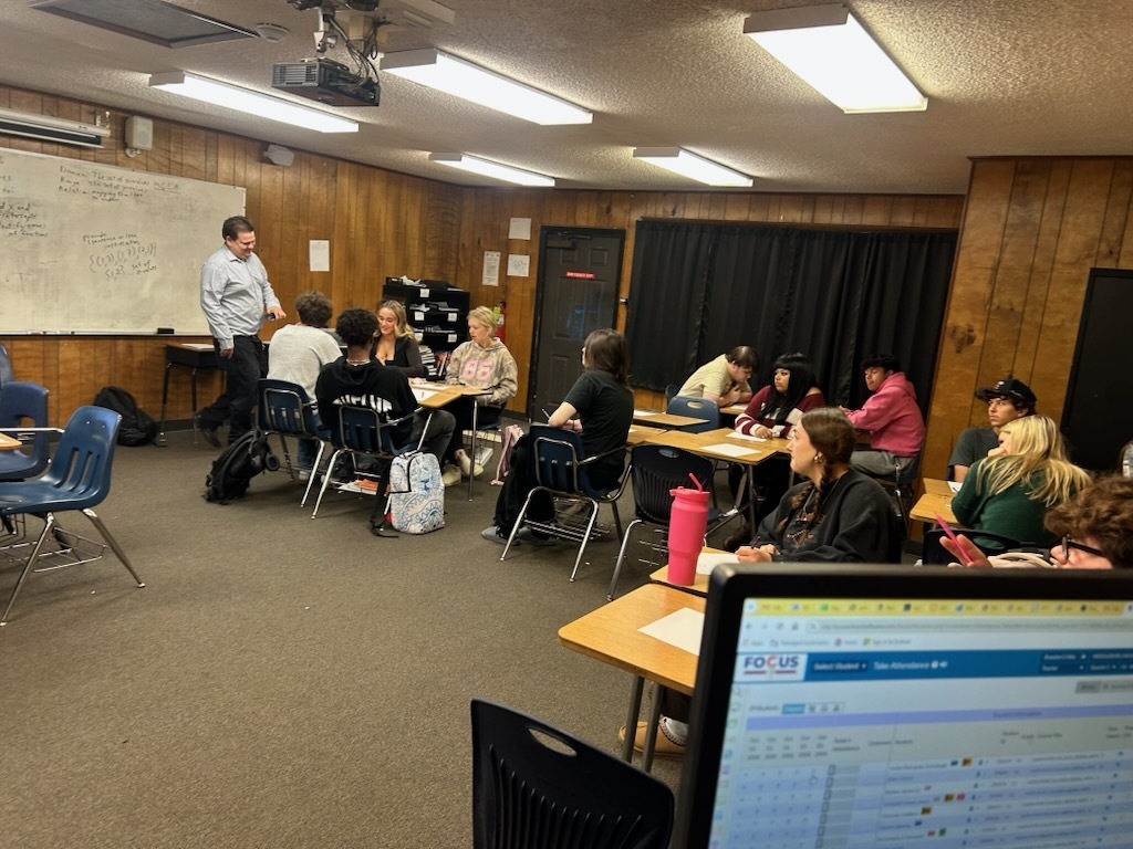 Classroom with tables, chairs, and a whiteboard. Students and a teacher gather around tables. Monitors and bags visible.