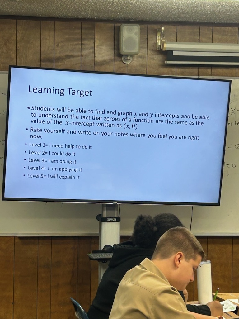 A classroom with wooden walls, a large screen showing a lesson on finding and graphing intercepts, and two students seated.