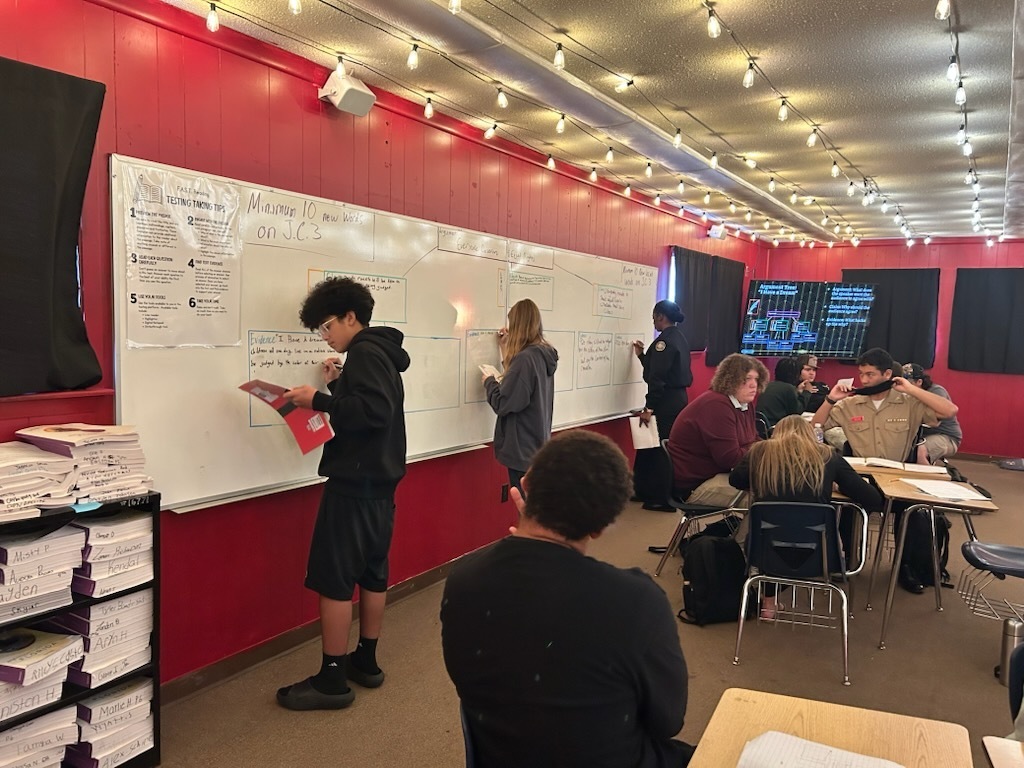 Students working in groups, some writing on whiteboards, others seated in chairs, with red walls, ceiling lights, and a dark backdrop.