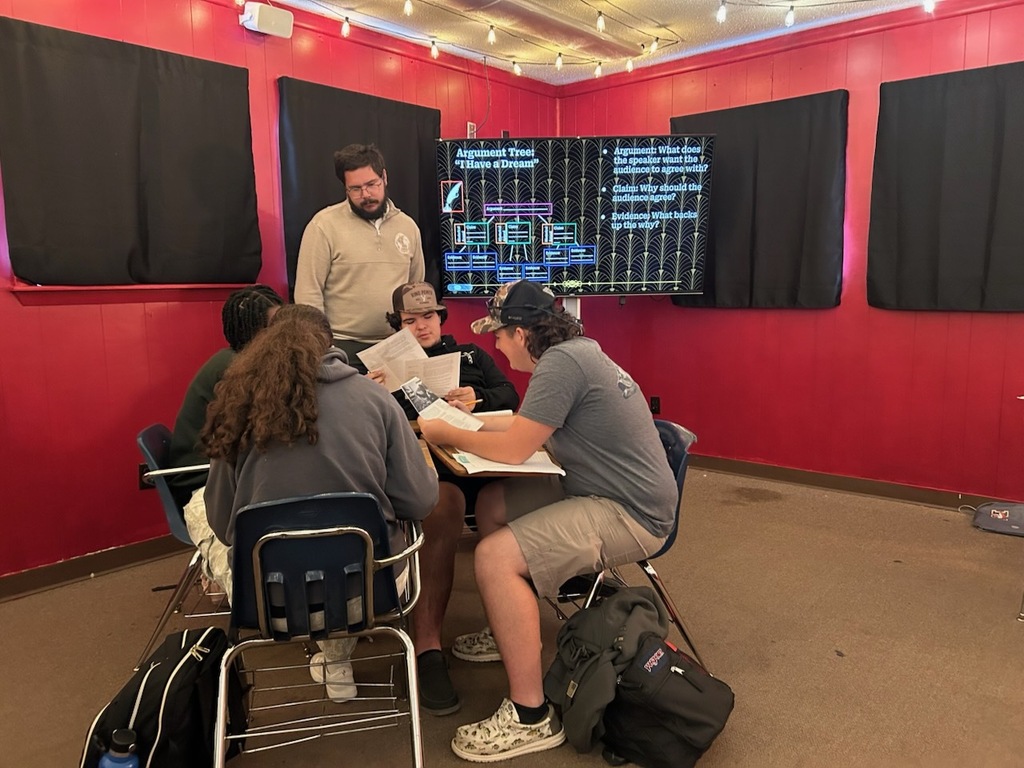 Five students sit in chairs around a table; a teacher stands nearby. A large screen displays text.