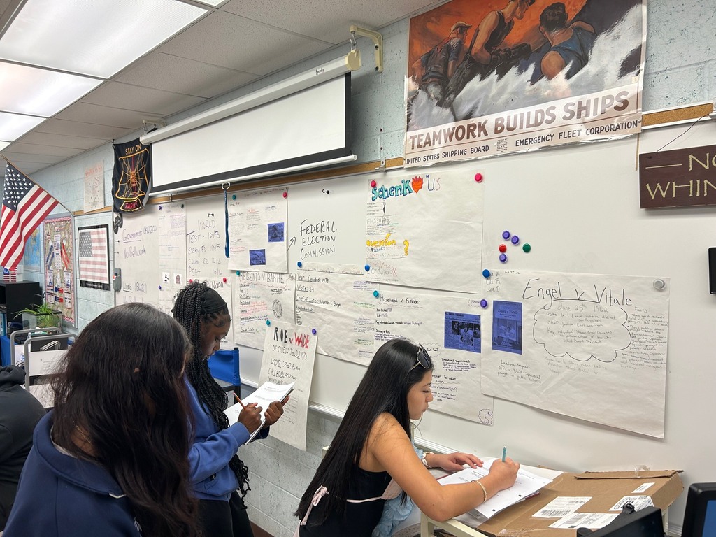 Three people at a desk in a classroom. A board with posters is behind them.