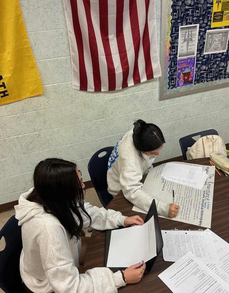 Two students in white hoodies are seated at a table. One holds a pen and paper, while the other works on a laptop.