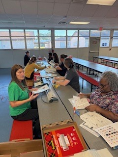 People sitting at tables with books and papers. One person holds a binder. Others wear glasses.