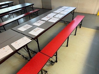 Long tables with papers on them and red benches in a large, empty room with white walls and a gray floor.