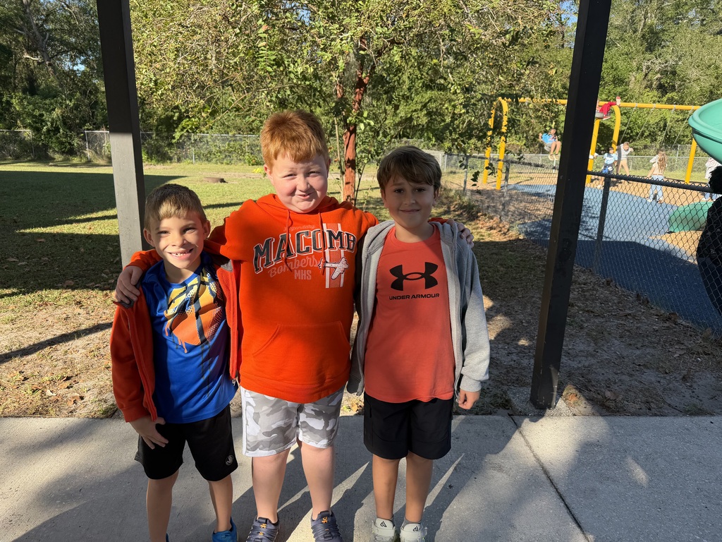 Three boys pose for a photo at an outdoor school playground, with trees and a slide visible.