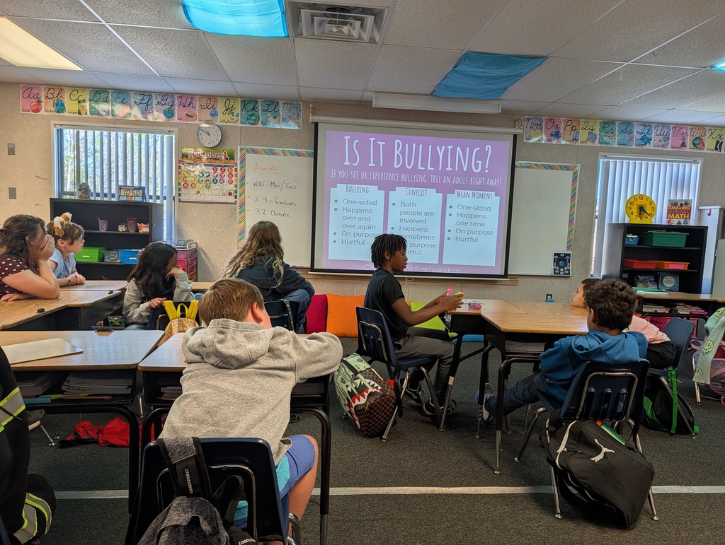 A classroom with students seated around desks. A projector screen displays text reading "Is It Bullying?"