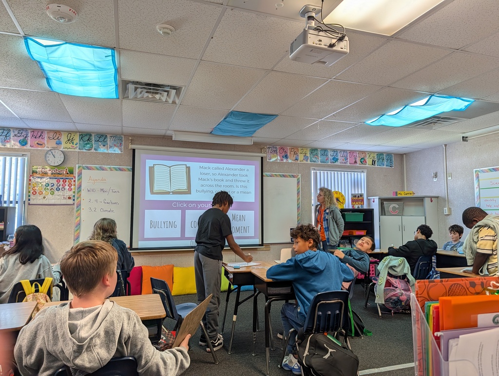 Classroom with students seated around tables; teacher at front with projector screen displaying an image; various materials on tables and shelves.