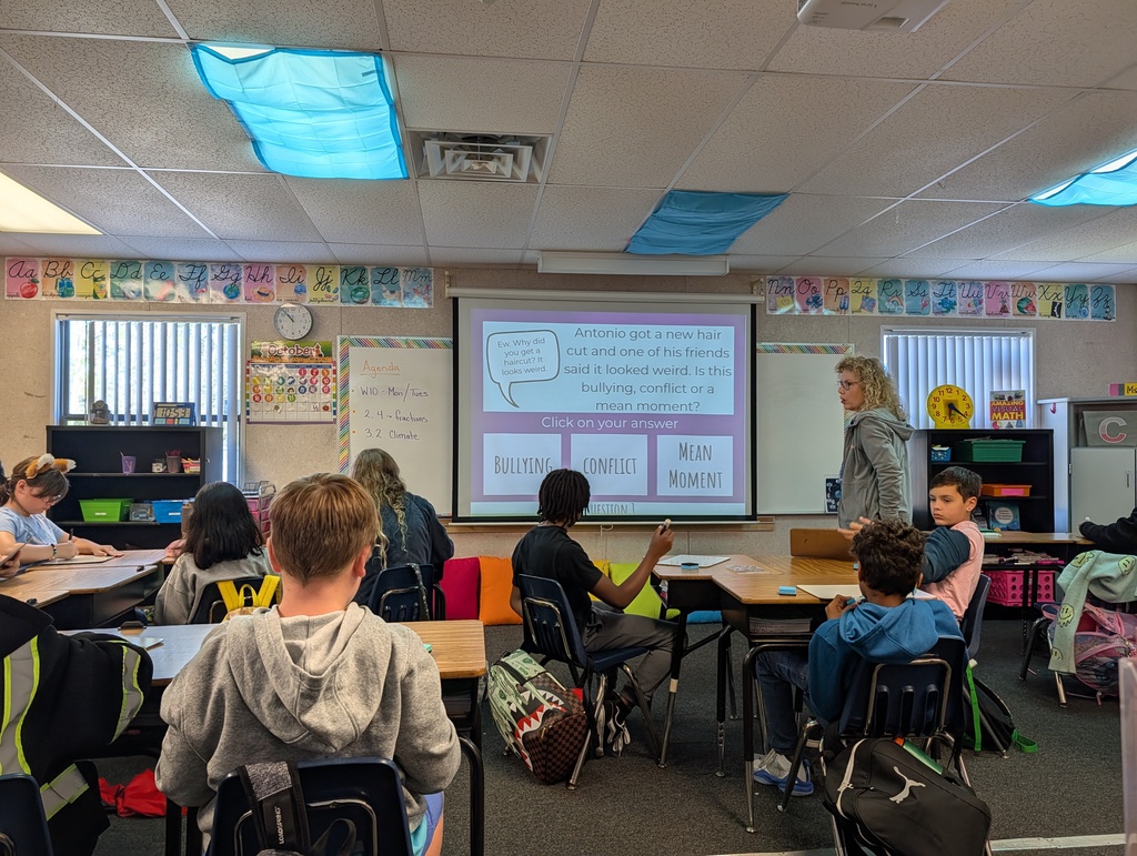 Students sit at desks in a classroom, with a teacher standing in front of a projector screen.